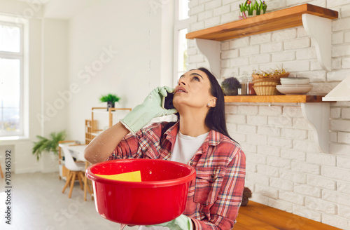 Having a roof leak at home. Worried young woman in rubber gloves holding red plastic basin, looking up at water dripping down from ruined ceiling and talking to repair and maintenance service on phone