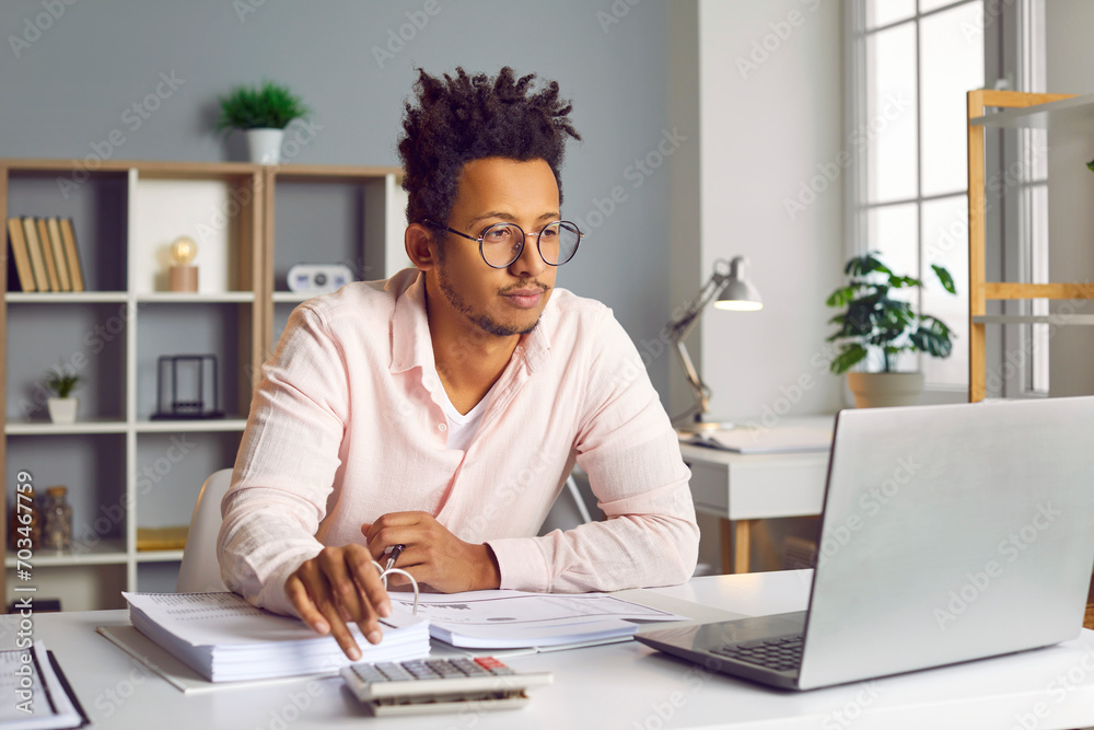 Young african american business man accountant working on a laptop at the office or at home, looking on a computer screen and making calculations, considering new projects, analyzing company. Young african american business man accountant working on a laptop at the office or at home, looking on a computer screen and making calculations, considering new projects, analyzing company.