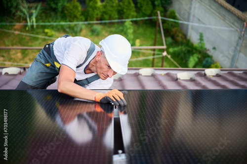 Worker building solar panel system on rooftop of house. Man engineer in helmet installing photovoltaic solar module outdoors. Alternative, green and renewable energy generation concept.