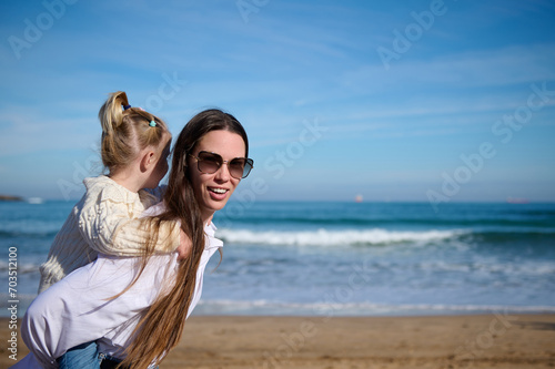 Cheerful young woman carrying her daughter on her back, giving her a piggyback ride while walking together on the beach.