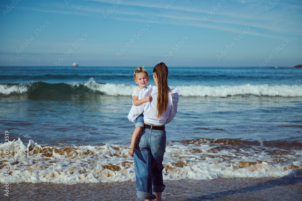 Rear view young mother carrying her daughter while walking barefoot with her cute child on the sandy beach Rear view young mother carrying her daughter while walking barefoot with her cute child on the sandy beach