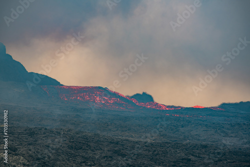 View across a hardened black grey lava field with lava and smoke erupting from a volcano in the distance - Iceland, Geldingadalir near Grindavik - june 2021