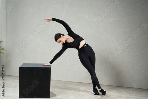 Graceful female athlete bending, while standing near cube, against grey wall. Side view of slim young woman in sportswear performing pose, while leaning on cube in gym. Concept of sporty lifestyle.