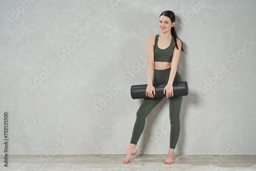 Pretty young woman in crop top posing with yoga mat in studio. Low angle view of attractive slim lady wearing activewear smiling to camera, while standing against grey wall, copy place. Sport concept.
