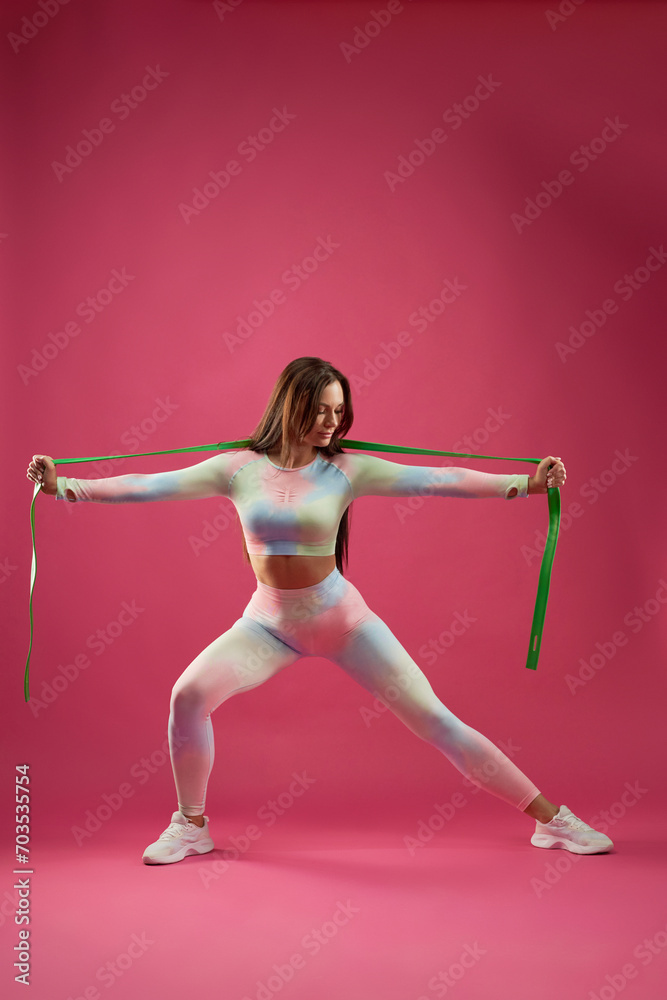 Strong female in sportswear stretching with rubber band during fitness workout indoors. Front view of motivated middle-aged woman doing lung with sport rope, against pink background. Sport concept.