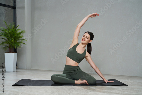 Young slender woman sitting in yoga pose with raised hand and meditating indoors. Graceful nice girl doing stretching, physical practice, exercising at home, against grey wall. Lifestyle concept.