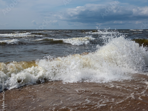 Close-up shot of big foamy waves in windy day of Baltic sea with brown sand on the coast and blue sky above. Seascape