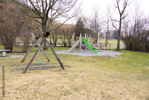 Large wooden children's playground with green swing and slide, meadow and gravel area, bare trees and bushes in the background, autumn, without people