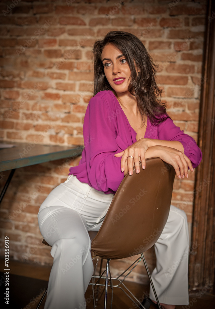 Beautiful young brunette woman in a purple blouse and white pants posing indoor against a brick wall. Portrait of a beautiful young brunette woman