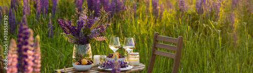Beautiful romantic outdoor wedding decor in field. Table decorated with purple lupines flowers. Wineglasses with white wine. Sunset, summer, golden hour. Perfect surprise date for loving couple banner