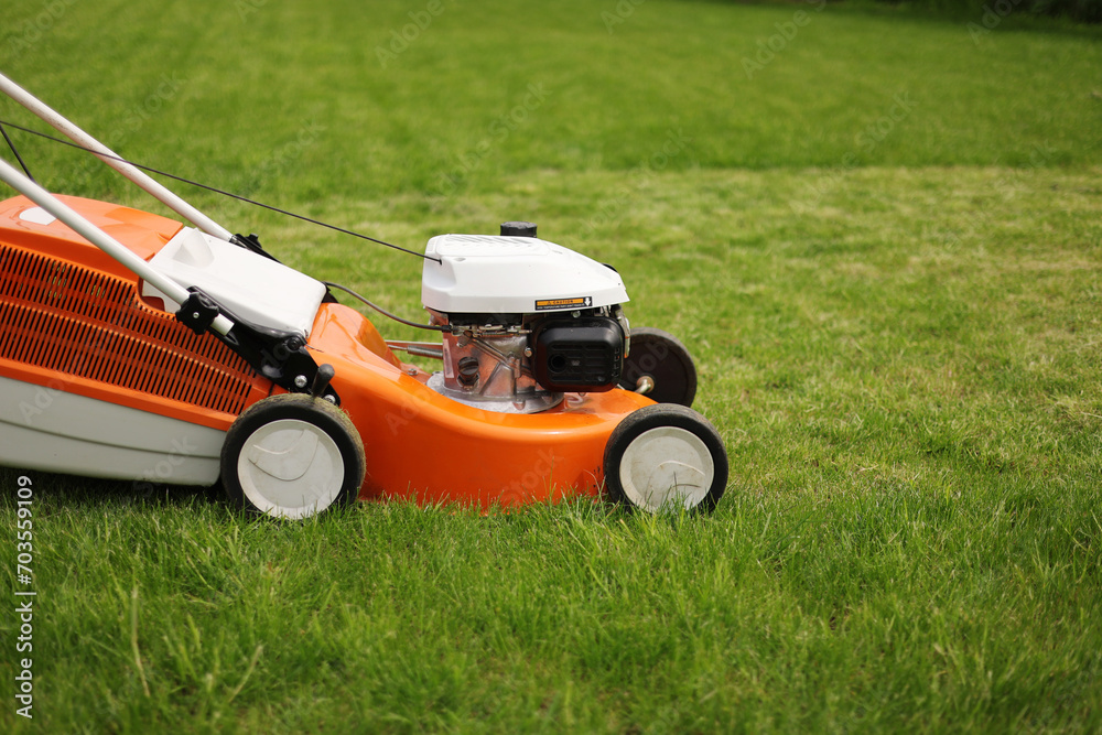 Fototapeta premium Close up orange-grey electric lawn mower on bright lush green lawn. Gardening work tools. Rotary lawn mower machine cut grass. Professional lawn care service. Place for text