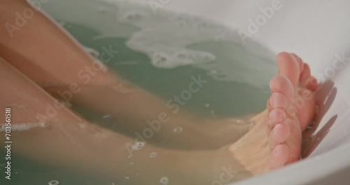 Anonymous female with neat feet splashing, while soaking in warm bathtub at home. Crop view of woman dipping feet in water, while having relaxing bath, in motion. Concept of relax, spa, body care. 