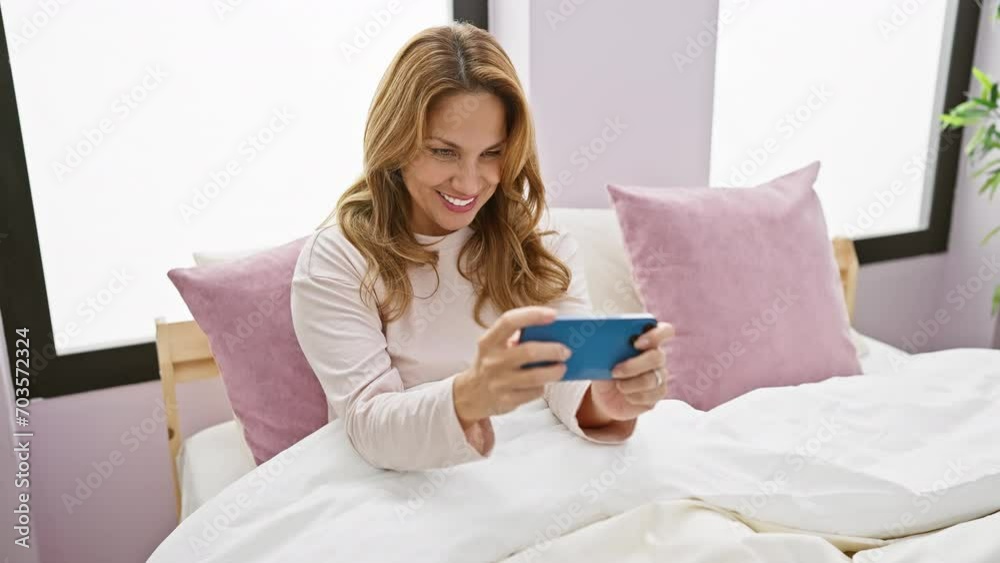 Smiling woman plays with smartphone on her bed in a bright bedroom, featuring modern pastel decor. Smiling woman plays with smartphone on her bed in a bright bedroom, featuring modern pastel decor.