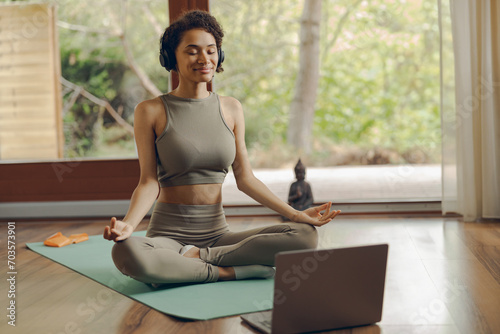 Woman in headphones practicing yoga and listen meditation at home sitting in lotus pose on mat