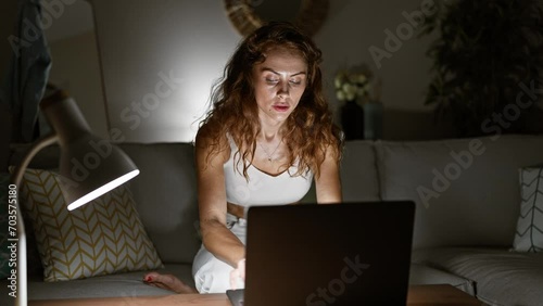 Caucasian woman working on laptop at home, illuminated by a lamp at night, evoking focus and dedication. photo