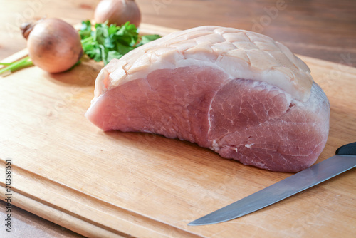 Raw salted roast pork on a wooden cutting board with kitchen knife, onions and herbs, cooking preparation for a festive Christmas ham with crust, copy space, selected focus