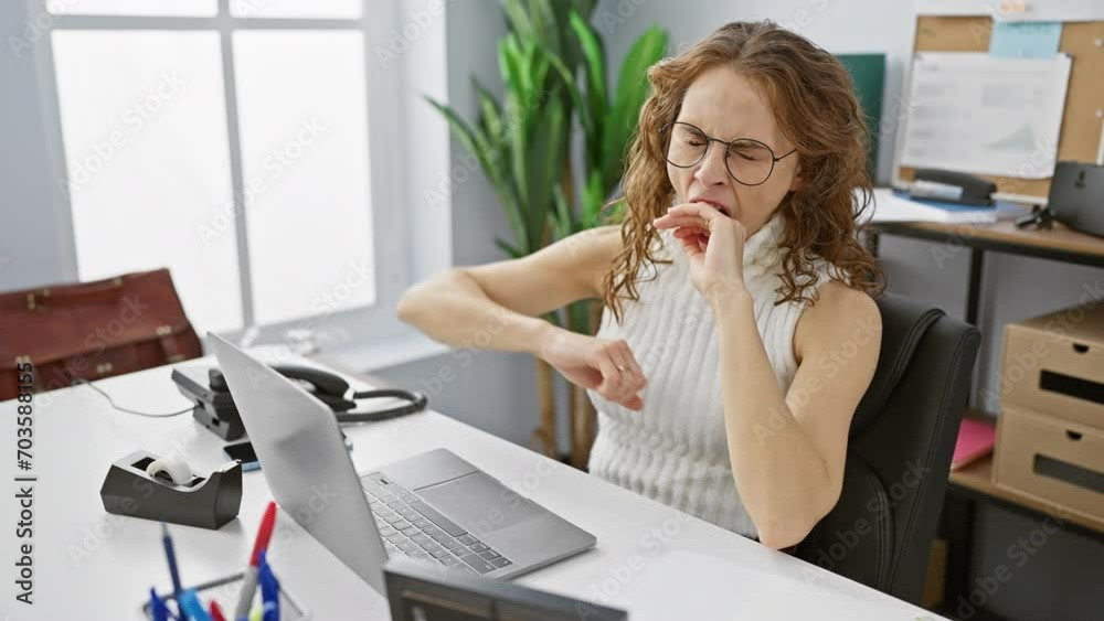 Overworked young woman, bored and yawning at the office, fighting sleepiness with hand over her mouth