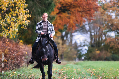 Young woman with red hair rides her horse along the edge of an autumn forest, photographed from the front. © RD-Fotografie