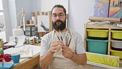 Hispanic man with beard holding paintbrushes in an artist's studio photo