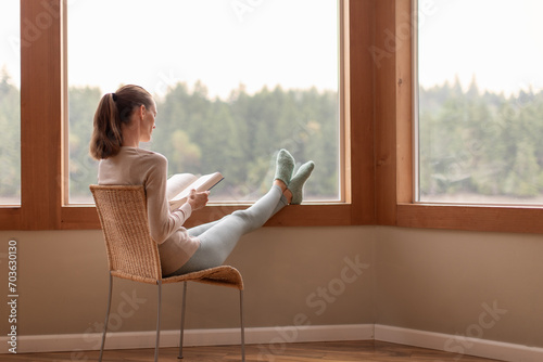 Young woman relaxing at home sitting next to window reading a book