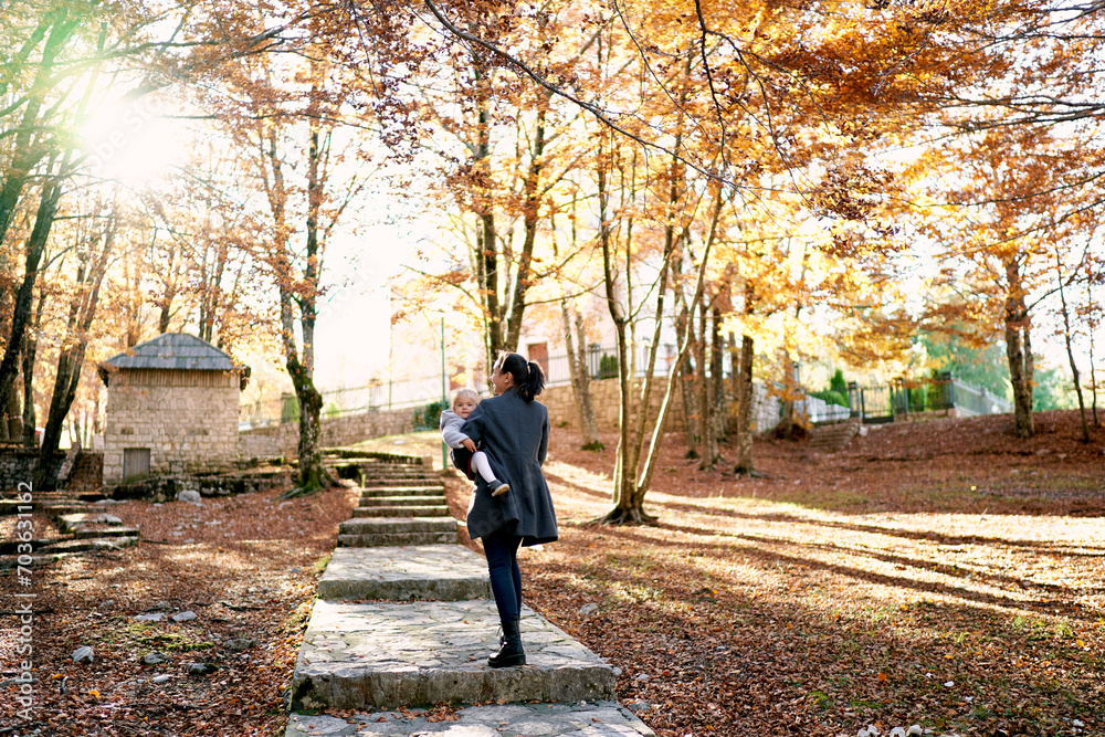 Mom with a little girl in her arms walks along a paved path with steps in an autumn park