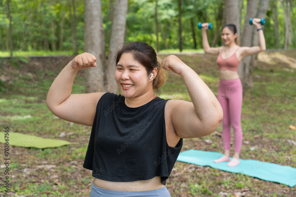 Happy smiling plus size Asian woman putting her arms up to show her arms' size while a skinny woman  holding small dumbbells  standing in the background at an outdoor park
