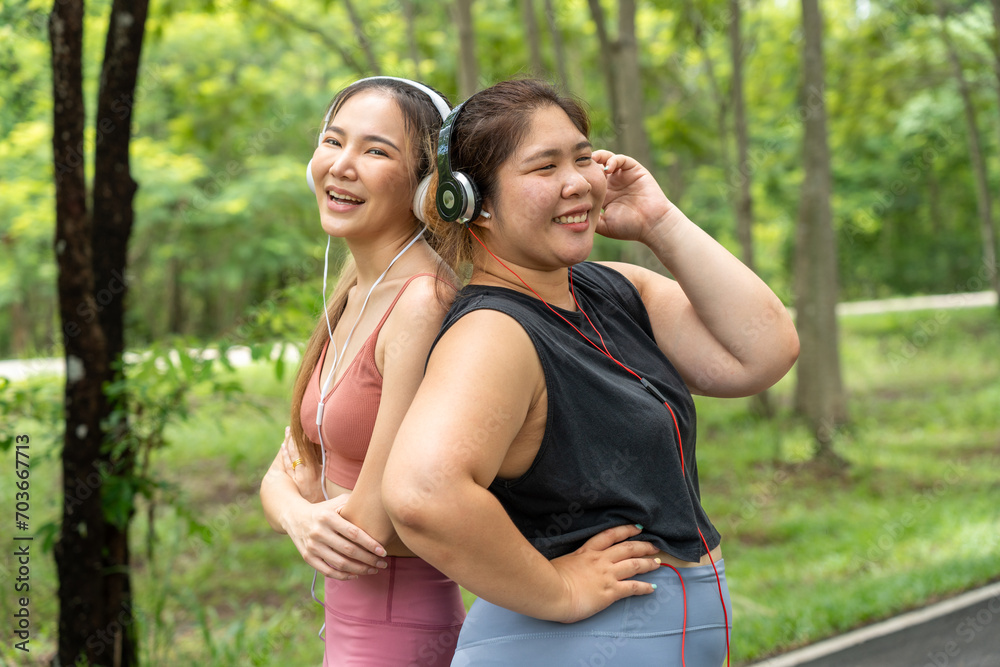 Two happy Asian female friends, one over size and one lean, standing with their back together listening to music from their headphones at a local park Two happy Asian female friends, one over size and one lean, standing with their back together listening to music from their headphones at a local park