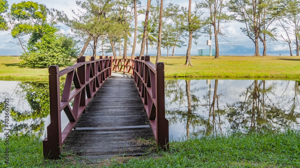 Obraz premium A pedestrian wooden bridge with railings spans the river. Green grass and trees grow on the lawn. The ocean is ahead. Clouds in the blue sky. A mirror image in calm water. Malaysia. Borneo. 