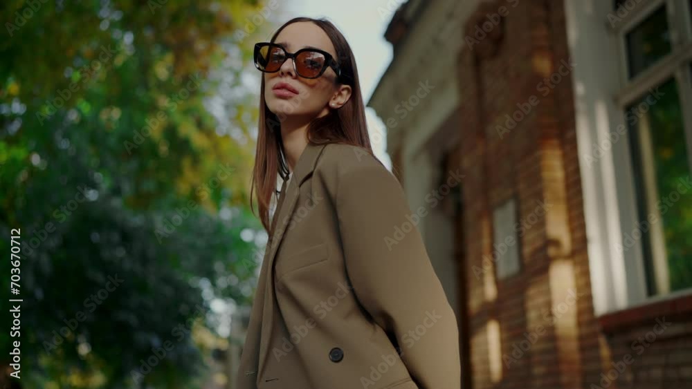 Beautiful girl in trendy jacket and sunglasses posing on camera outside. Young female in stylish clothes and glasses walking on city street.