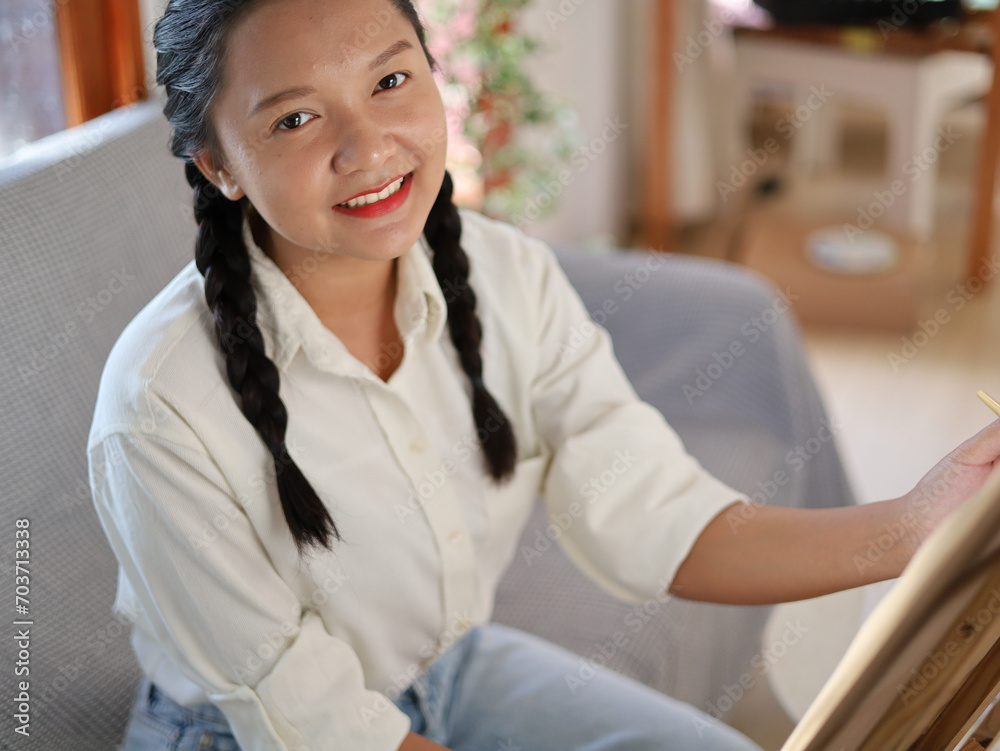 Happy young girl siting on sofa and drawing with a paint palette and a paintbrush at home,painting artist.