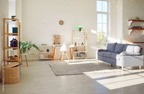 Simple interior design of a new clean modern apartment. No one at home, in a cozy bright sunny living room with white walls, gray sofa, wooden shelves, green plants, warm rug and laminate flooring 