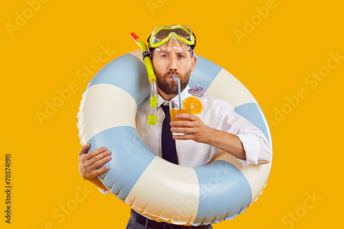 Portrait of a funny bearded man in scuba glasses drinking orange juice cocktail holding inflatable rubber ring isolated on yellow studio background. Summer holiday trip and vacation concept photo