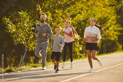 Fototapeta Naklejka Na Ścianę i Meble -  Sporty family jogging together. Happy mother, father and children in sportswear running on asphalt road lined with green trees on sunny summer morning. Outdoor sports workout concept