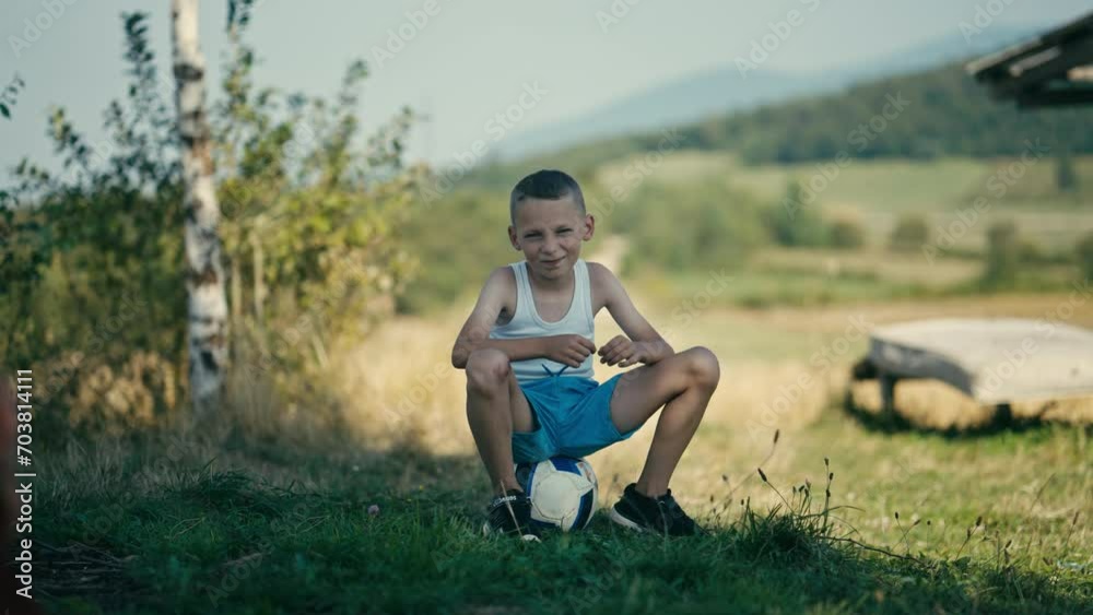Blond Countryside Boy Smiling While Sitting on a Ball and Tearing the Grass. Concept of Future Athlete, Aspiring Footballer, Summer Olympics, UEFA EURO, FIFA World Cup Blond Countryside Boy Smiling While Sitting on a Ball and Tearing the Grass. Concept of Future Athlete, Aspiring Footballer, Summer Olympics, UEFA EURO, FIFA World Cup