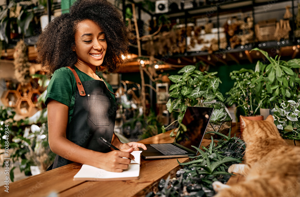 Eco friendly market store. Cheerful female gardener with curly hair smiling to red cat lying on desk at atmospheric flower shop. Black woman in green apron noting data from modern laptop on clipboard.