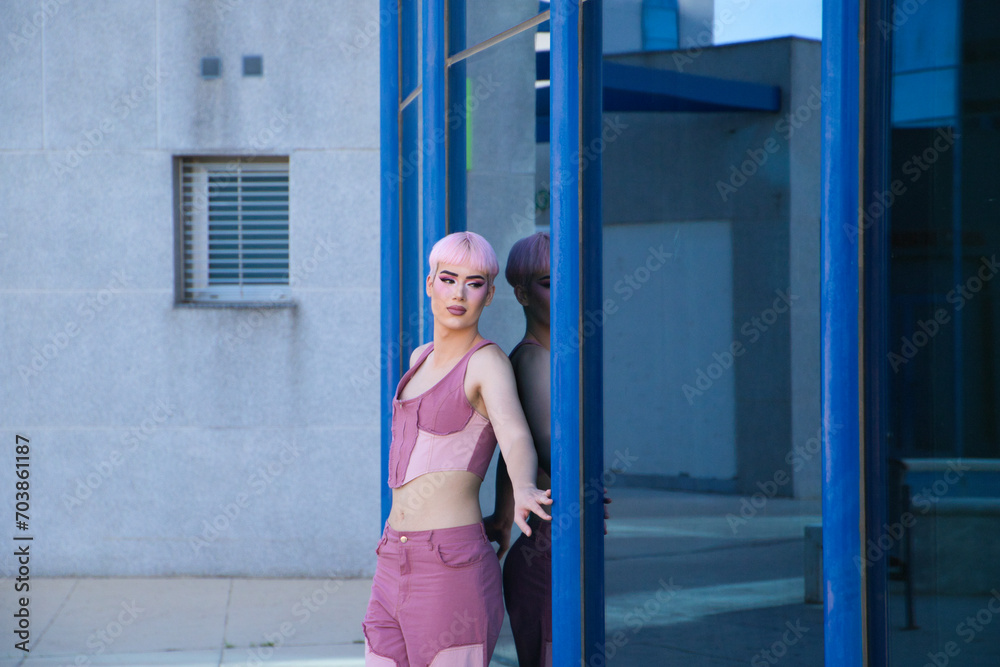 Young gay guy with pink hair and make-up leans against a glass and mirrored office building. The boy is dressed in pink. Concept of equality and LGBTQ rights. Young gay guy with pink hair and make-up leans against a glass and mirrored office building. The boy is dressed in pink. Concept of equality and LGBTQ rights.