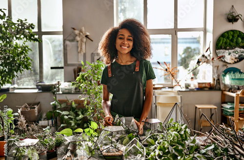 Floristic work at shop. Smiling store worker standing by wooden desk full of plants and gardening equipment. African american woman filling glass polyhedron florarium with stones and succulent.