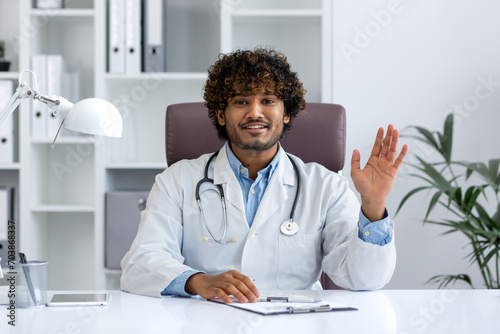 Webcam view, online video call consultation, young Indian doctor in white medical coat looking at camera, waving hand greeting to patient, doctor working remotely inside clinic office with laptop. photo