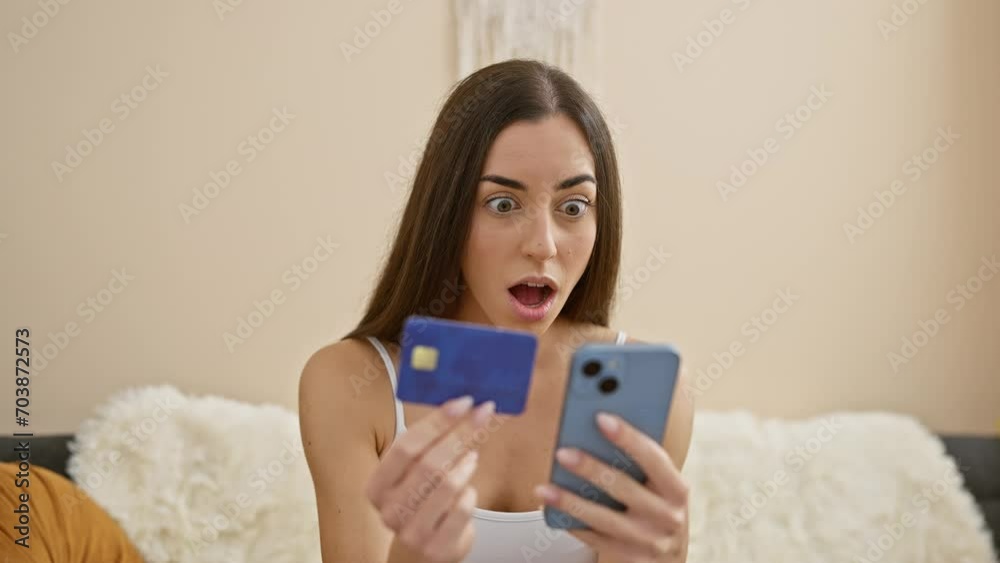 Surprised young hispanic woman sits on her home sofa, smartphone in hand, beautiful and intense as she engages in online shopping with her credit card. Surprised young hispanic woman sits on her home sofa, smartphone in hand, beautiful and intense as she engages in online shopping with her credit card.