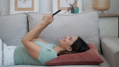 Video of pretty young woman sending messages with her mobile phone while lying down on sofa at home. photo