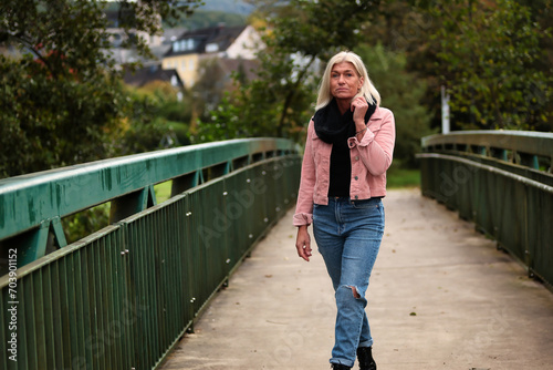 Woman blonde in her fifties with a pink denim jacket, blue jeans with a ripped black sweater and a black scarf, portrait on a bridge. © RD-Fotografie