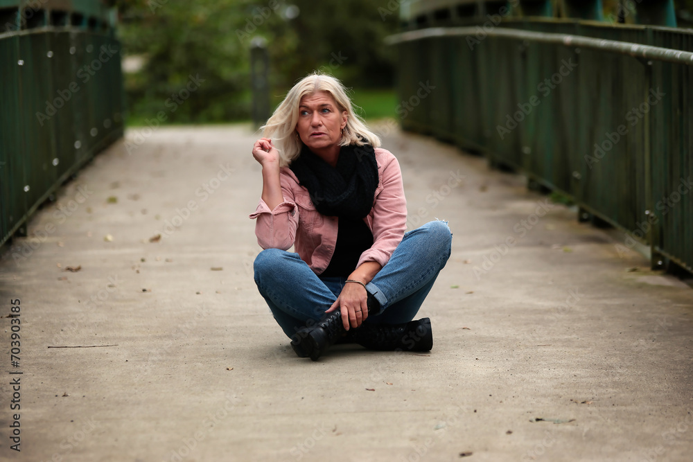 Woman blonde in her fifties with a pink denim jacket, blue jeans with a ripped black sweater and a black scarf, portrait on a bridge.