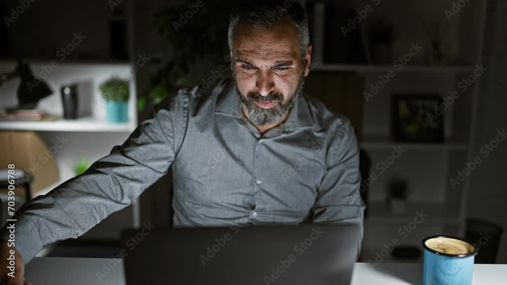 Mature bearded man working late in a dark office, focused on laptop screen with coffee beside him. Mature bearded man working late in a dark office, focused on laptop screen with coffee beside him.