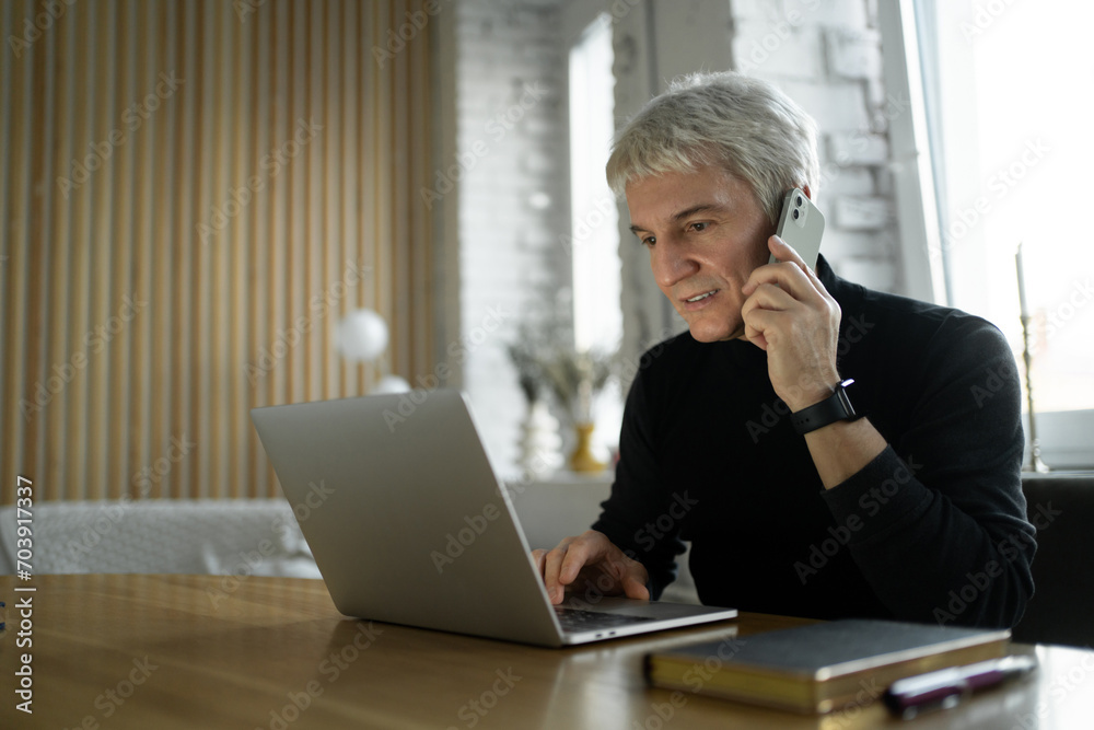 Focused senior professional multitasking with a laptop and phone in a stylish office setting. Focused senior professional multitasking with a laptop and phone in a stylish office setting.