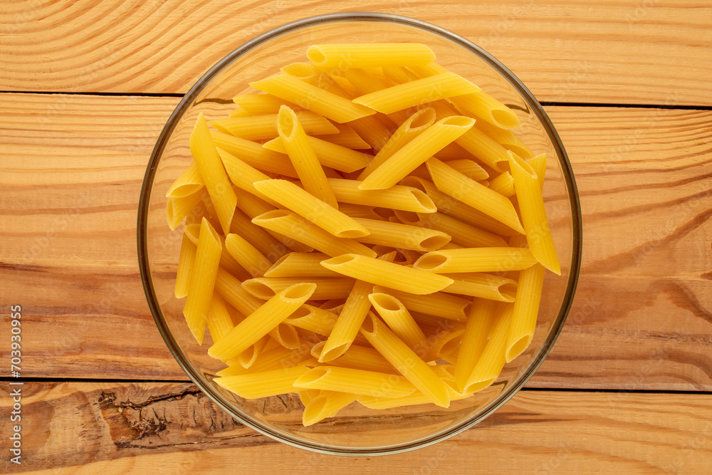 Raw pasta, penne, in glass plate on wooden table, macro, top view.