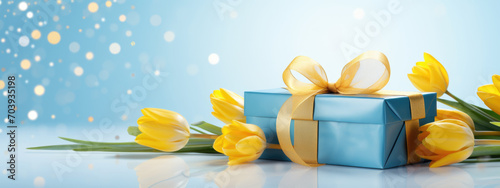Bright yellow tulips and elegantly wrapped gifts adorned with blue and golden ribbons against a soft blue background with light bokeh and golden glitter on the surface. © MP Studio
