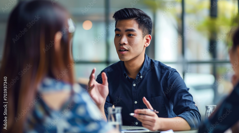 Man in a conversation, gesturing with his hands while speaking to an out-of-focus person across the table, in a well-lit indoor environment suggestive of a professional meeting. - obrazy, fototapety, plakaty 