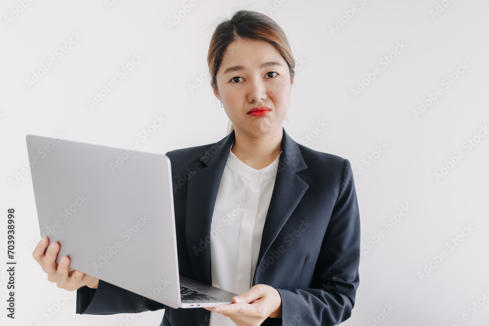 Unhappy asian Thai woman wear blazer suit, looking at camera with sad face, dislike holding laptop and looking at computer, standing over white background wall. Unhappy asian Thai woman wear blazer suit, looking at camera with sad face, dislike holding laptop and looking at computer, standing over white background wall.
