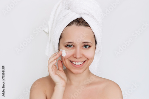 Young caucasian attractive smiling woman with towel on her head after shower touching face with hand applying a moisturizing cream on the skin isolated on a white background. Cosmetology and beauty