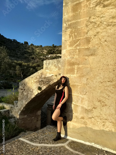 Vintage pin-up, donned in black and red, poses by a medieval tower on Gargano with a bold slit and captivating smile, enhancing the allure amid the historic setting.